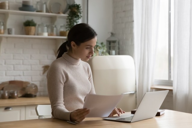 Concentrated woman holding documents and working on laptop.