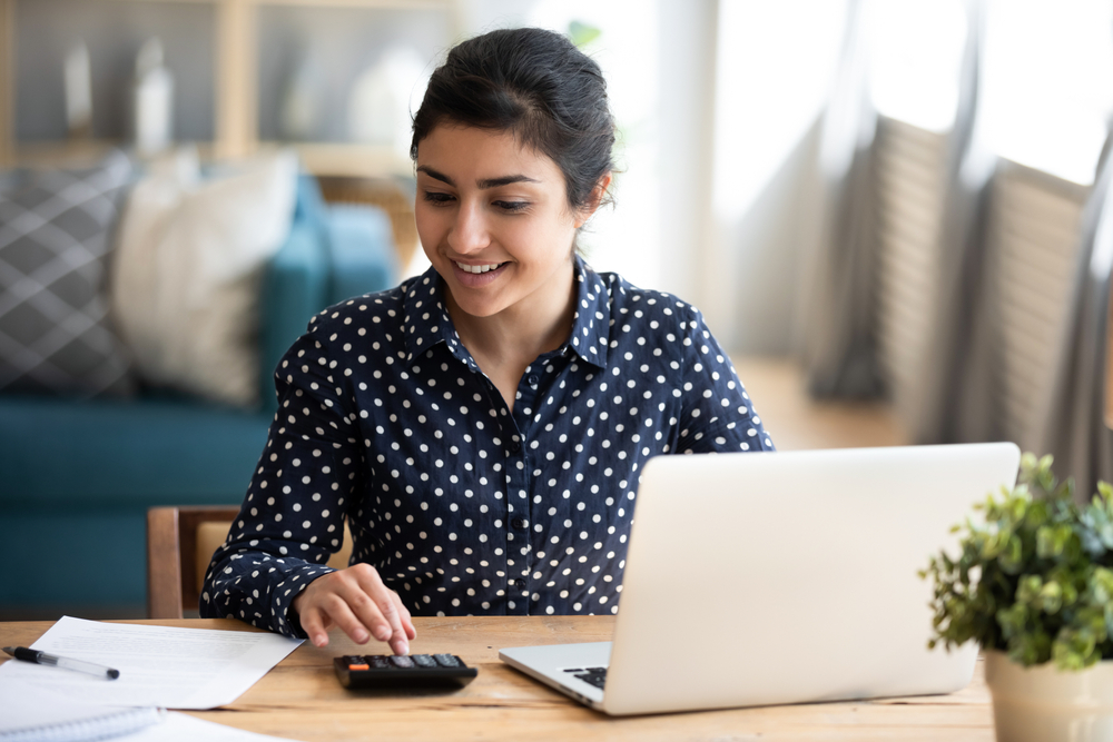 Woman sitting at a table using her laptop and a calculator.