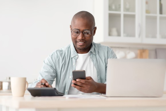 Image of man sitting at the kitchen table with a laptop, his phone, and a calculator.