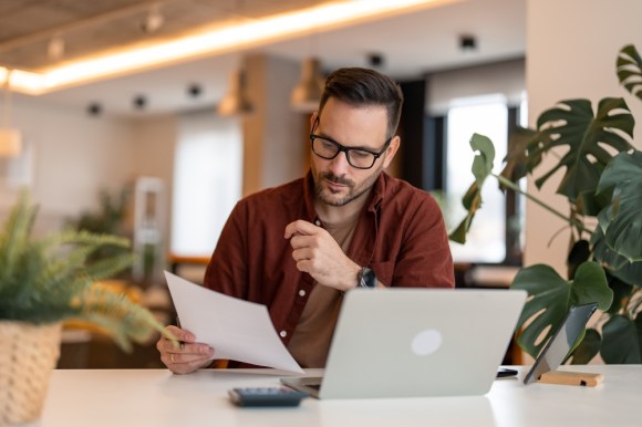 Man sitting in a cafe in front of a laptop holding a document.