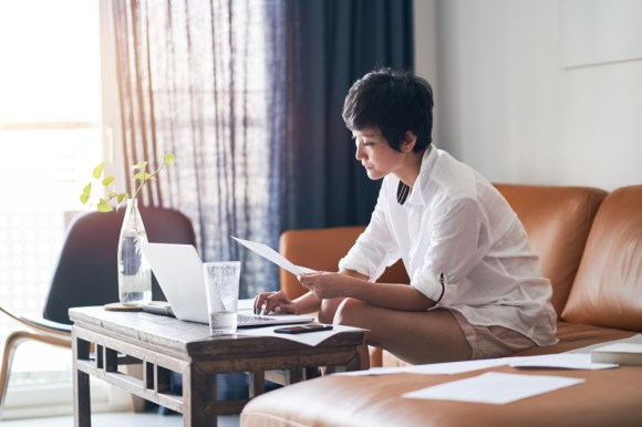 Asian woman sitting on her couch using her laptop with documents spread around her.
