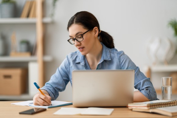 Woman in home office sitting in front of a table with a laptop open and writing in a notebook. 