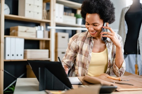 Black woman sitting at a desk talking on the phone and taking notes.