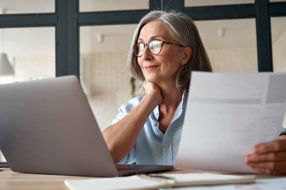 Elderly woman holding a document in one hand and looking at her laptop.