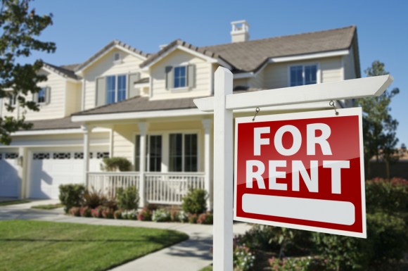 Left-facing red “For Rent” real estate sign in front of a yellow house.