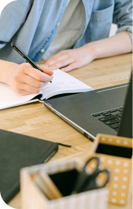 Photo of a person writing in a notebook at a desk in front of a laptop