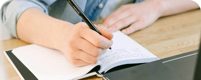 Photo of a person writing in a notebook at a desk in front of a laptop