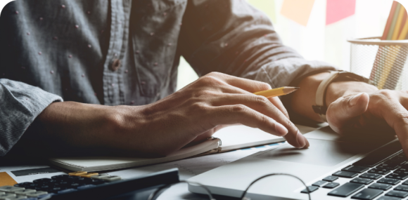 Photo of a user's hands using a laptop and holding a pencil near a notebook