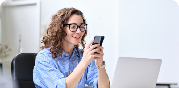 Photo of a woman smiling at her phone while sitting at her laptop