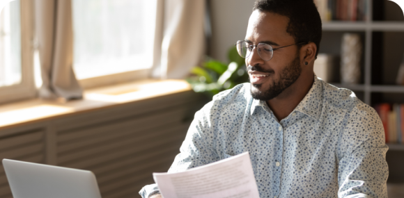 Photo of a man smiling at his laptop while holding papers